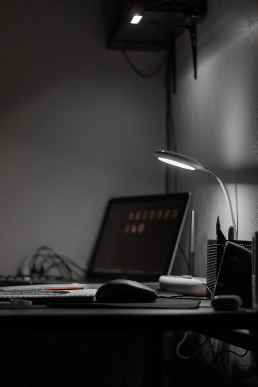 A desk with a computer monitor, a lamp, and various office supplies, including pens and a mouse, in a dimly lit environment.