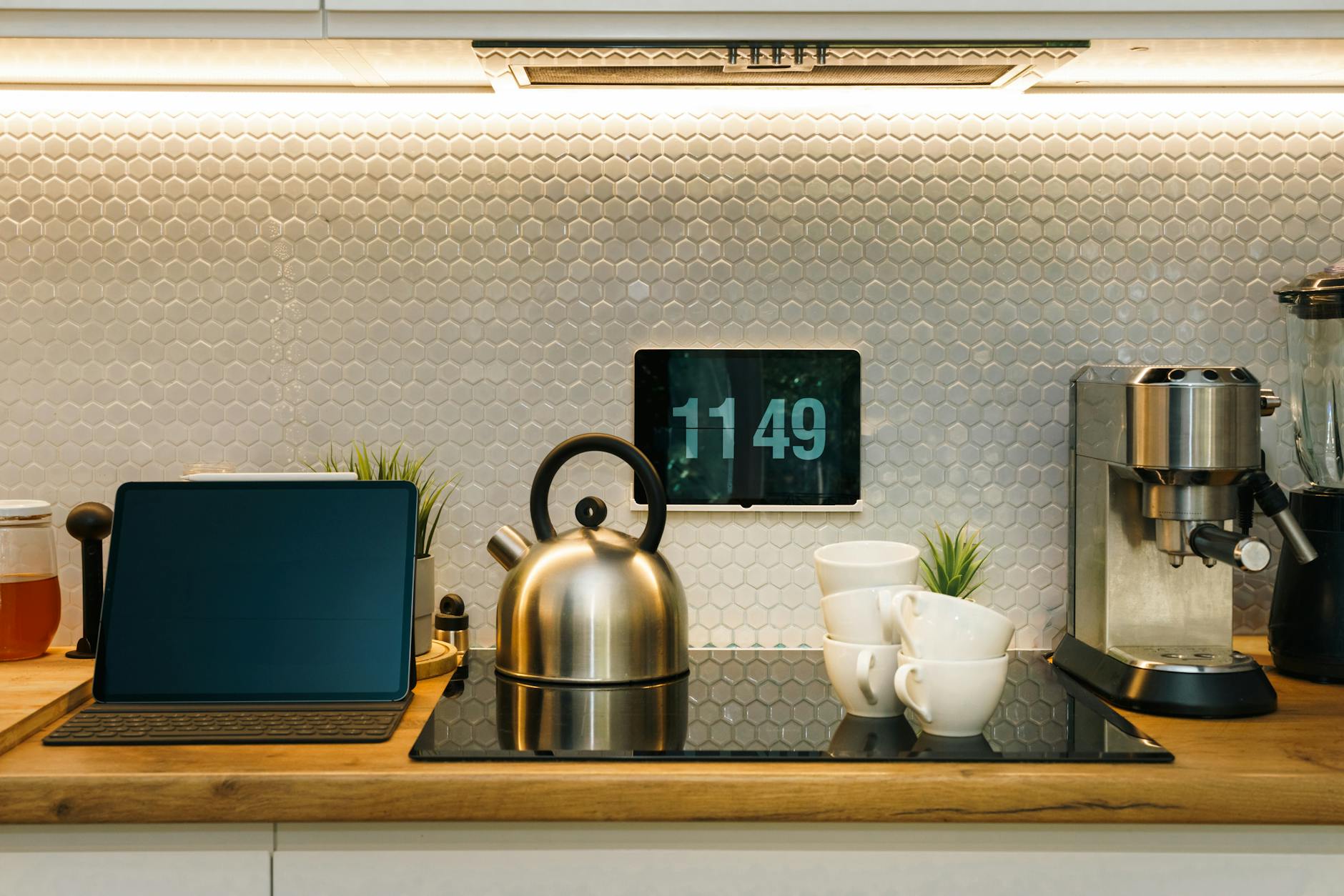 A modern kitchen counter featuring a sleek kettle, several white cups, a digital clock displaying the time, and a coffee machine, set against a textured hexagonal tile backsplash.