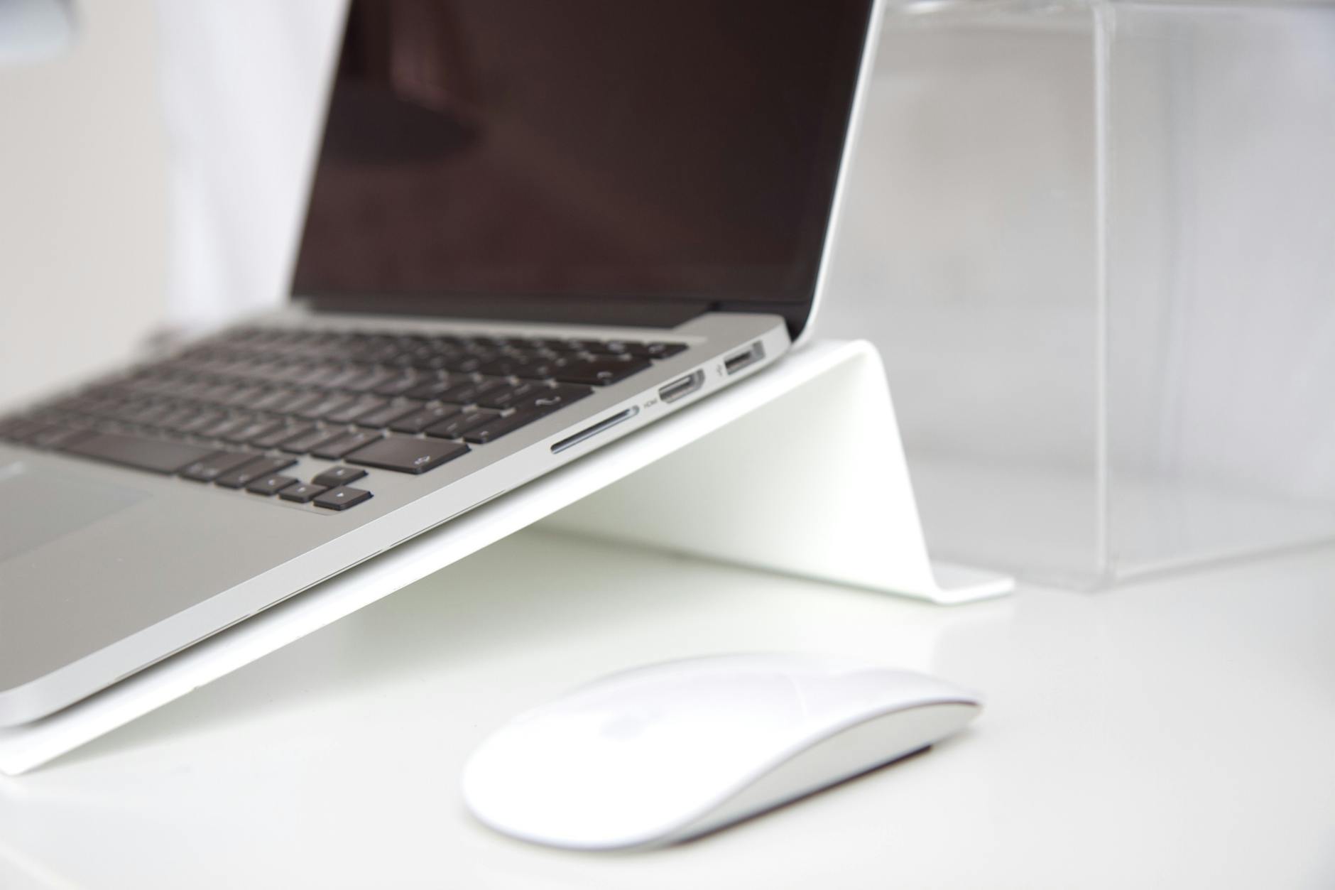 A laptop positioned on a white stand, with a mouse placed nearby on a desk.